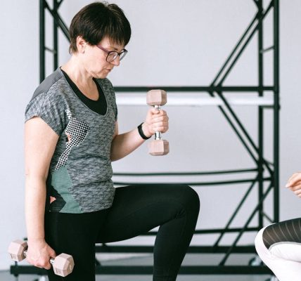 Elderly woman lifting dumbbells with guidance from a trainer indoors.