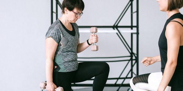 Elderly woman lifting dumbbells with guidance from a trainer indoors.